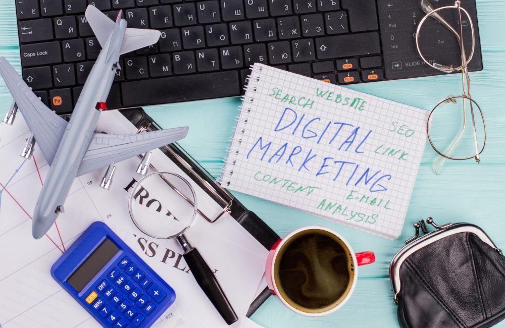 Digital marketing on notepad and various office supplies. Flat lay composition with keyboard, coffee and other office related objects on blue background.